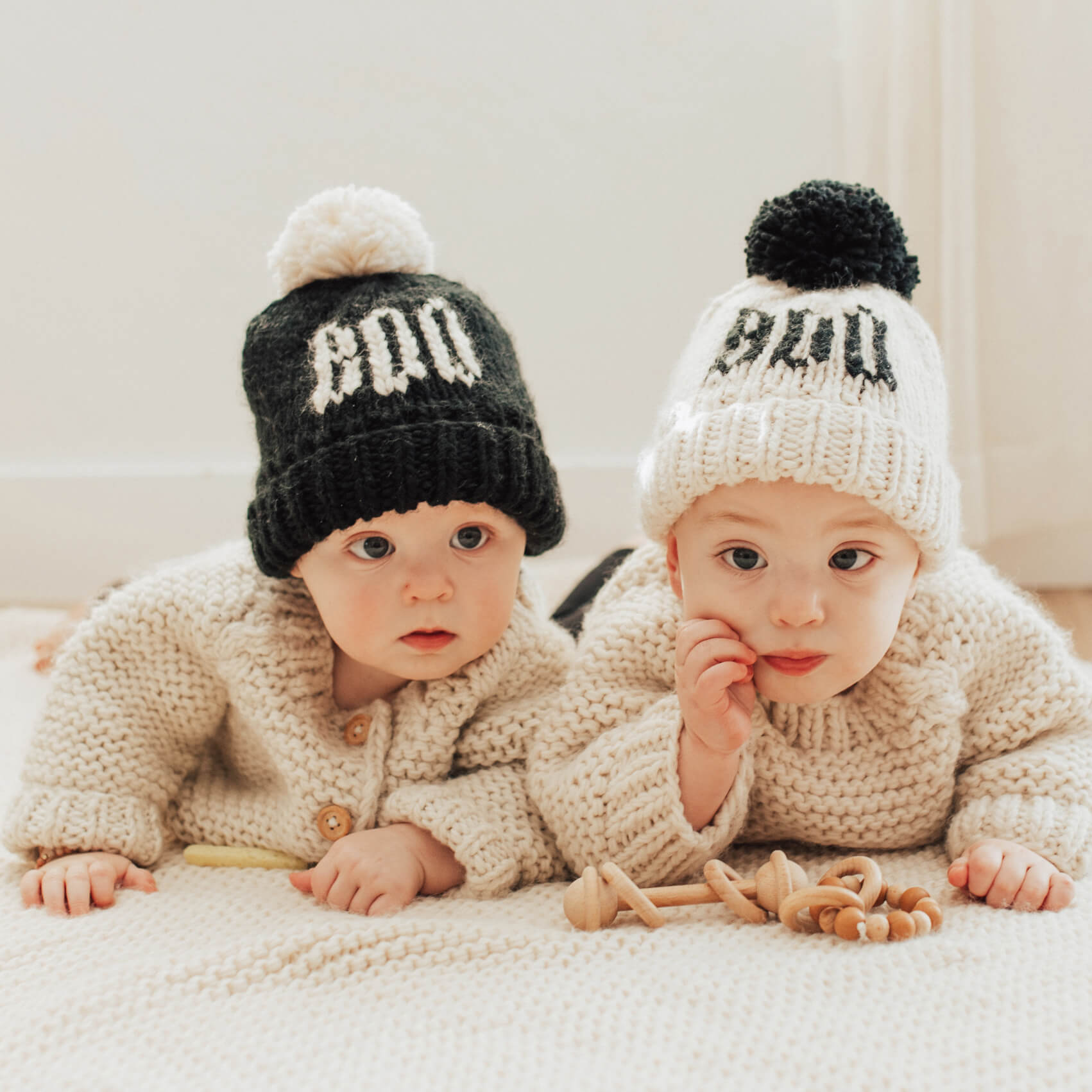 two babies wearing knit hats. one is black with natural BOO on the front and the other is natural with black BOO